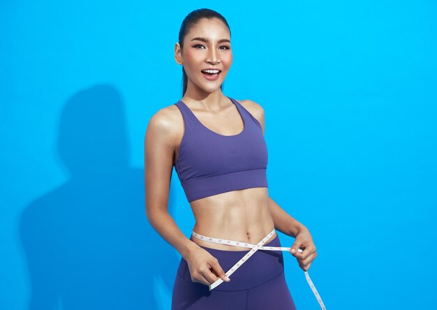 Joven y hermosa deportista asiática sosteniendo cinta métrica alrededor de su cintura sobre fondo azul.