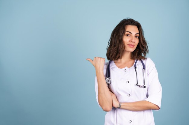 Una joven y hermosa doctora con una bata de laboratorio de fondo azul sonríe y señala con el dedo a la izquierda en un espacio vacío