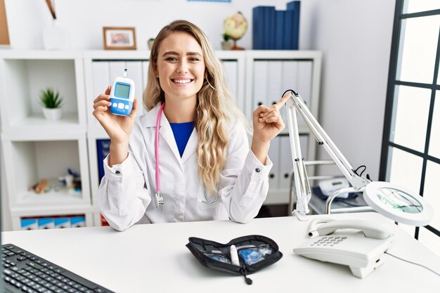 Joven y hermosa doctora sosteniendo un medidor de glucosa sonriendo feliz señalando con la mano y el dedo a un lado