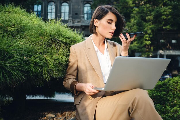 Joven hermosa y elegante mujer de negocios que trabaja en una computadora portátil grabando mensajes de voz al aire libre