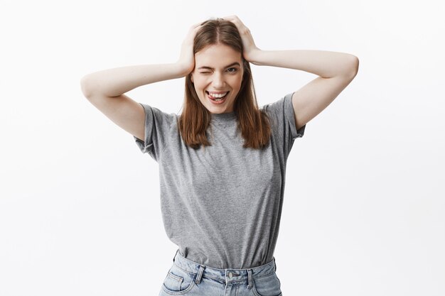 Joven hermosa estudiante caucásica con cabello oscuro y ojos marrones en ropa gris casual, guiñando un ojo, sonriendo con dientes y mostrando la lengua, siendo feliz.