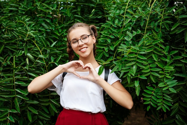 Joven hermosa estudiante en gafas mostrando corazón, posando sobre las hojas al aire libre.