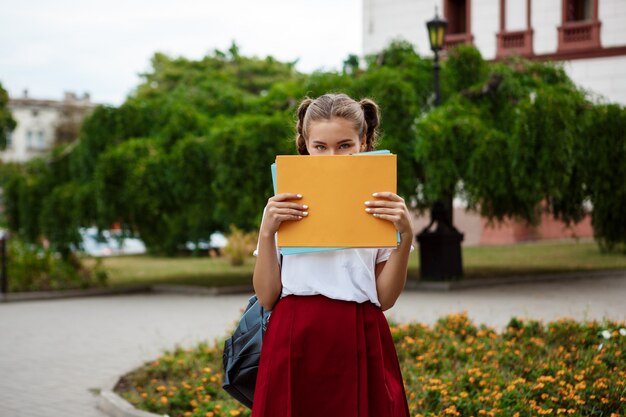 Joven hermosa estudiante sosteniendo carpetas delante de la cara, al aire libre