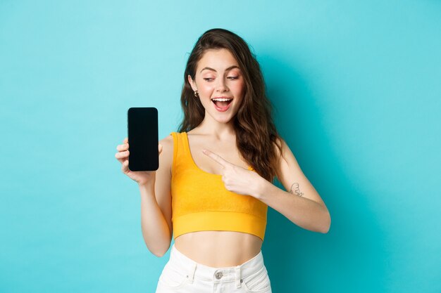 Joven hermosa feliz, apuntando y mirando la pantalla vacía del teléfono inteligente, demostrar publicidad en la pantalla del móvil, de pie contra el fondo azul.