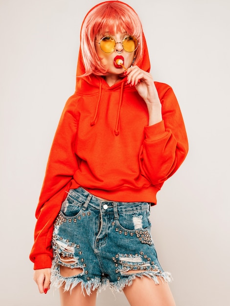 Joven hermosa hipster chica mala en moda rojo verano rojo con capucha y arete en la nariz. Mujer despreocupada sexy posando en estudio sobre fondo gris en peluca. Modelo caliente lamiendo dulces de azúcar redondos
