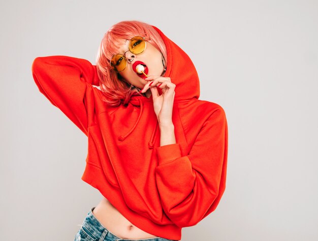 Joven hermosa hipster chica mala en moda rojo verano rojo con capucha y arete en la nariz. Mujer despreocupada sexy posando en estudio sobre fondo gris en peluca. Modelo caliente lamiendo dulces de azúcar redondos