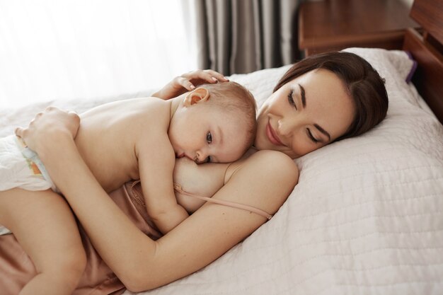 Joven hermosa madre feliz amamantando abrazando a su bebé acostado en cama en casa.