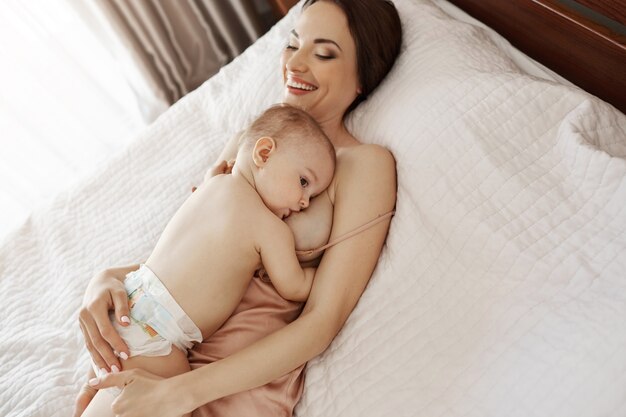 Joven hermosa madre feliz amamantando abrazando a su bebé acostado en cama en casa.
