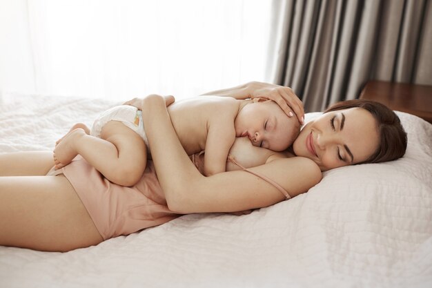 Joven hermosa madre feliz amamantando abrazando a su bebé acostado en la cama en su casa