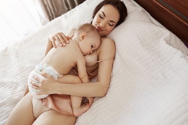 Foto gratuita joven hermosa madre feliz amamantando abrazando a su hijo acostado en la cama en su casa