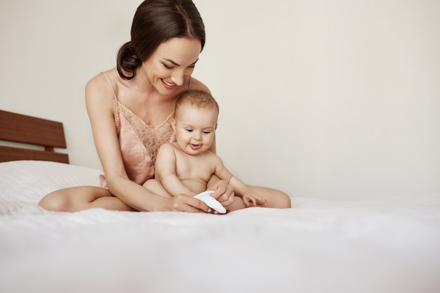 Joven hermosa madre feliz en ropa de dormir y su bebé recién nacido sentado en la cama en la mañana sonriendo jugando juntos.