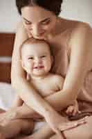 Foto gratuita joven hermosa madre feliz en ropa de dormir y su bebé recién nacido sentado en la cama sonriendo jugando juntos en casa.