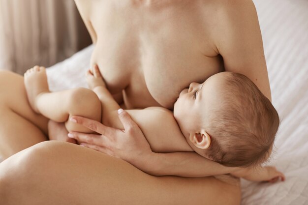Joven hermosa mamá desnuda amamantando abrazando a su bebé recién nacido sonriendo sentado en la cama en su casa
