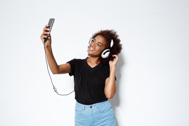 Joven hermosa mujer africana en auriculares haciendo selfie, sonriendo sobre la pared blanca