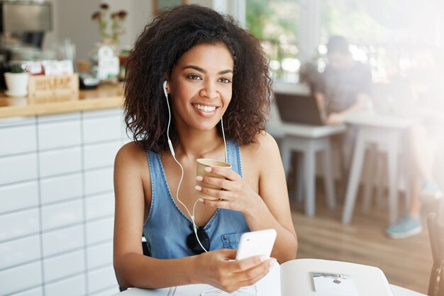 Joven hermosa mujer africana estudiante escuchando música en auriculares sonriendo sentado a la mesa con los libros en el café.