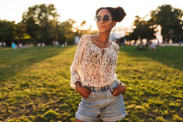 Joven hermosa mujer africana con gafas de sol mirando soñadoramente a la cámara mientras pasa tiempo en el parque de la ciudad