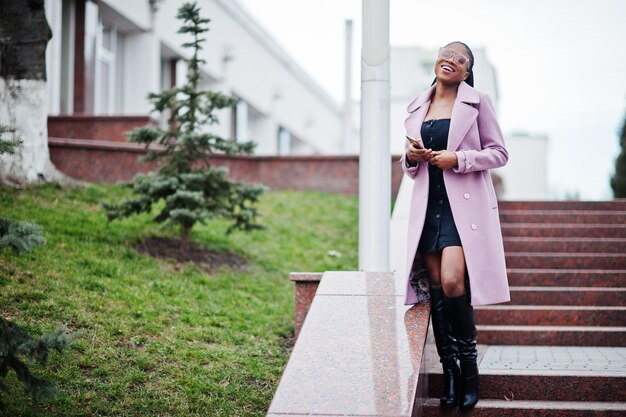 Joven y hermosa mujer afroamericana con estilo en la calle contra las escaleras usando anteojos de abrigo de moda con el teléfono móvil en las manos