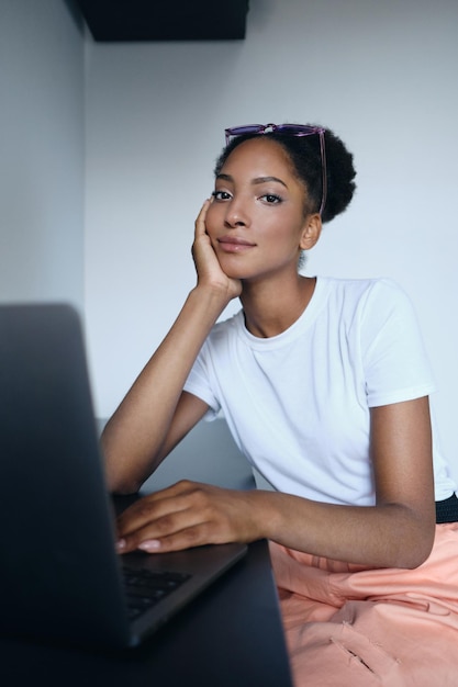 Joven y hermosa mujer afroamericana informal que trabaja en una laptop mirando cuidadosamente a la cámara en un moderno espacio de trabajo conjunto