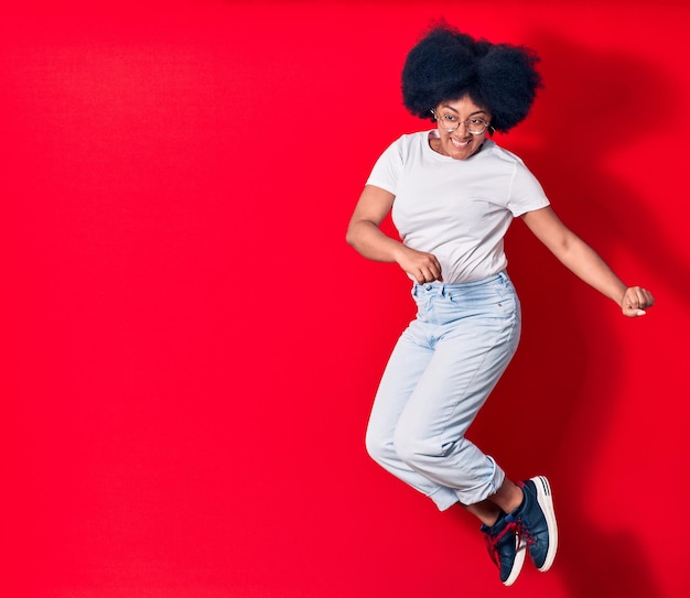 Foto gratuita joven hermosa mujer afroamericana con ropa informal y gafas sonriendo feliz. saltando con una sonrisa en la cara celebrando con los puños sobre un fondo rojo aislado