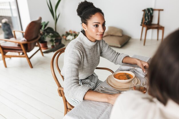 Foto gratuita joven y hermosa mujer afroamericana sonriente con cabello oscuro y rizado sentada en la mesa mirando soñadoramente a un lado mientras pasa tiempo con un amigo en un acogedor café