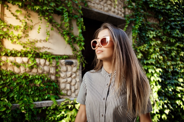 Joven hermosa mujer alegre en gafas de sol caminando por la ciudad, sonriendo.