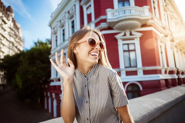 Joven hermosa mujer alegre en gafas de sol caminando por la ciudad, sonriendo.