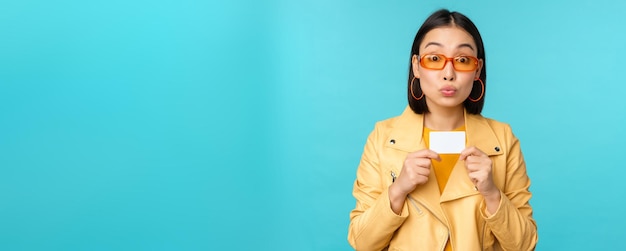 Joven hermosa mujer asiática mostrando tarjeta de crédito sonriendo eligiendo banco de pie sobre fondo azul.