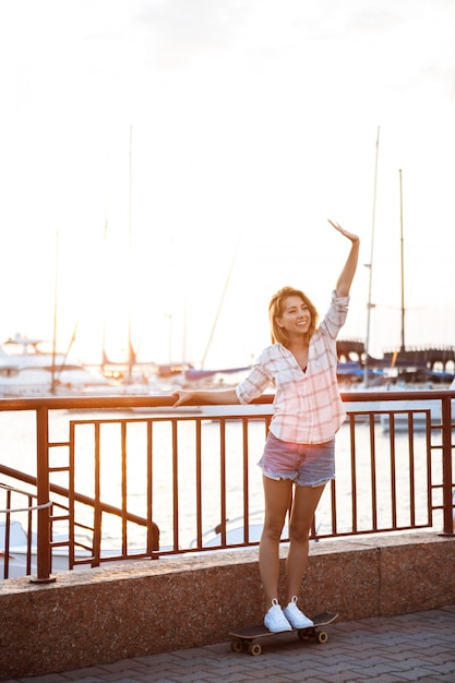 Joven hermosa mujer caminando en la playa, sonriendo, saludo, skate.