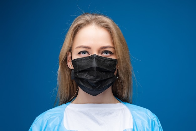Joven hermosa mujer caucásica en bata médica azul y con máscara médica blanca en su rostro se ve en la cámara