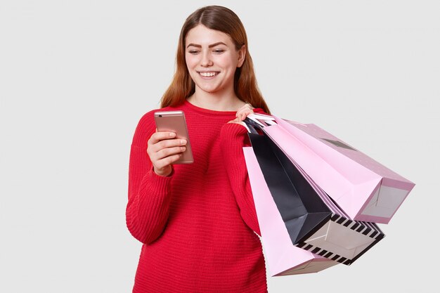 Joven hermosa mujer caucásica de moda tiene bolsas de compras en una mano y teléfono inteligente en otro aislado en blanco