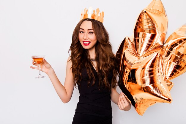 Joven hermosa mujer celebrando en vestido negro sonrisa y posando con cóctel en mano y globos de pureza.