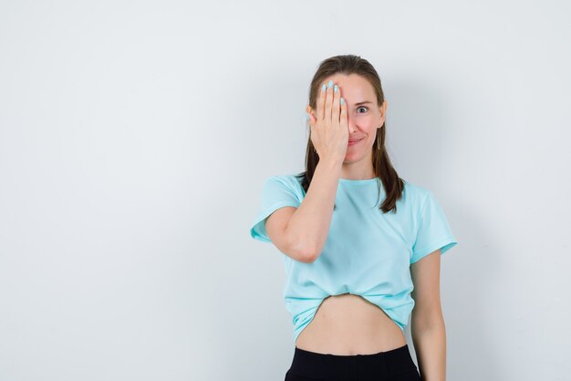 Joven hermosa mujer cubriendo los ojos con la mano en camiseta y mirando alegre. vista frontal.