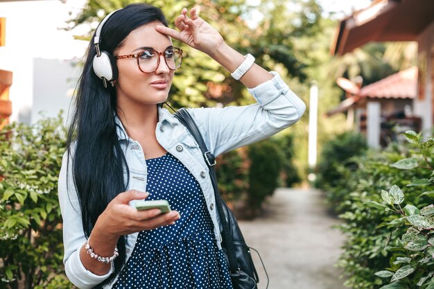 Joven hermosa mujer elegante con smartphone, auriculares, gafas, verano, traje de mezclilla vintage, sonriente, feliz, positivo