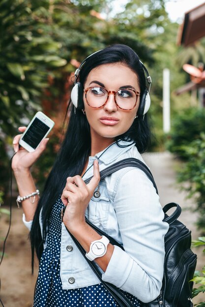 Joven hermosa mujer elegante con smartphone, auriculares, gafas, verano, traje de mezclilla vintage, sonriente, feliz, positivo