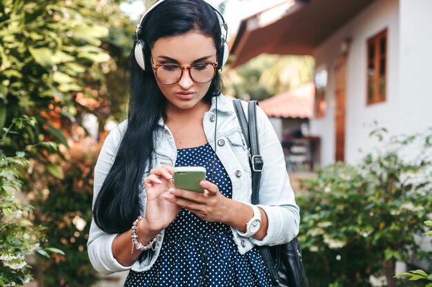 Joven hermosa mujer elegante con smartphone, auriculares, gafas, verano, traje de mezclilla vintage, sonriente, feliz, positivo