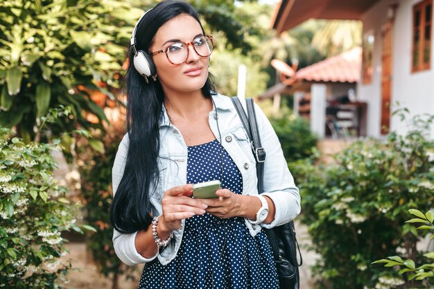 Joven hermosa mujer elegante con smartphone, auriculares, gafas, verano, traje de mezclilla vintage, sonriente, feliz, positivo