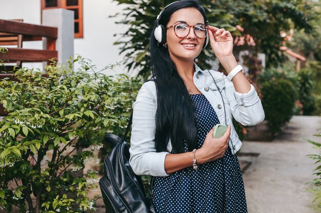 Joven hermosa mujer elegante con smartphone, auriculares, gafas, verano, traje de mezclilla vintage, sonriente, feliz, positivo