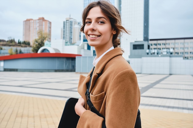 Joven hermosa mujer elegante sonriente con abrigo mirando alegremente a la cámara caminando por las calles de la ciudad