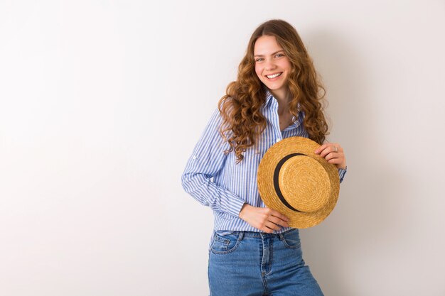 Joven hermosa mujer elegante en traje de estilo de verano posando en la pared blanca con sombrero de paja