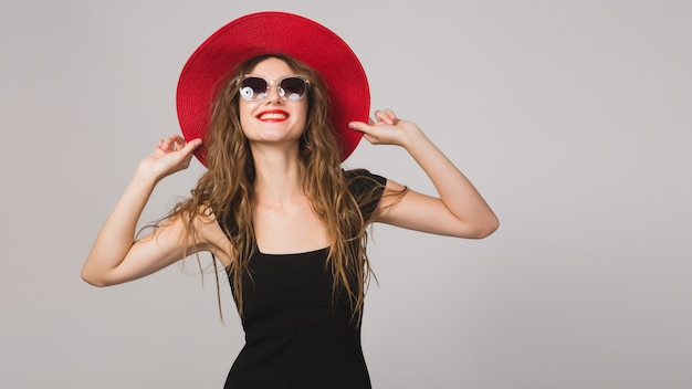 Joven hermosa mujer elegante en vestido negro, sombrero rojo, gafas de sol, lápiz labial rojo, feliz, sonriente, sexy, elegante