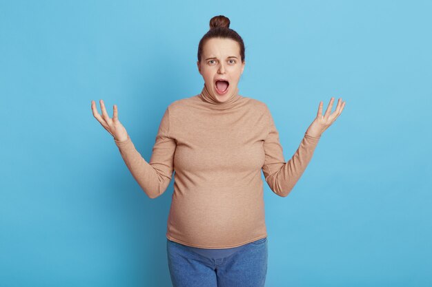 Joven hermosa mujer embarazada con moño, vestida con suéter casual y jeans, niña esperando un bebé, posando aislada sobre una pared azul, luciendo enojada, estresada y nerviosa, levantando las manos, gritando