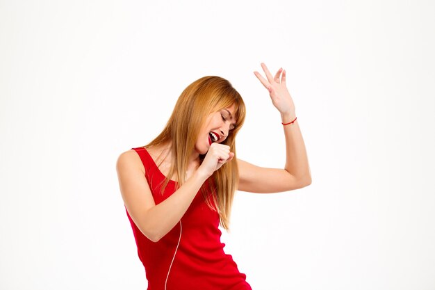 Joven hermosa mujer escuchando música en auriculares y cantando sobre pared blanca