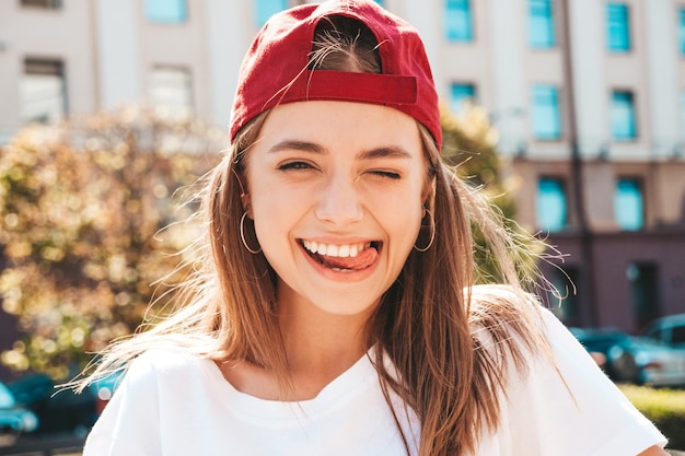 Joven hermosa mujer hipster sonriente en camiseta blanca de moda de verano Mujer sexy despreocupada posando en el fondo de la calle con gorra al atardecer Modelo positivo al aire libre Alegre y feliz Guiñando un ojo