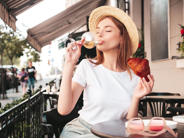 Joven hermosa mujer hipster sonriente en ropa de verano de moda Mujer despreocupada posando en el café de la terraza en la calle Modelo positivo bebiendo vino blanco Disfrutando de las vacaciones Comiendo croissant Con sombrero