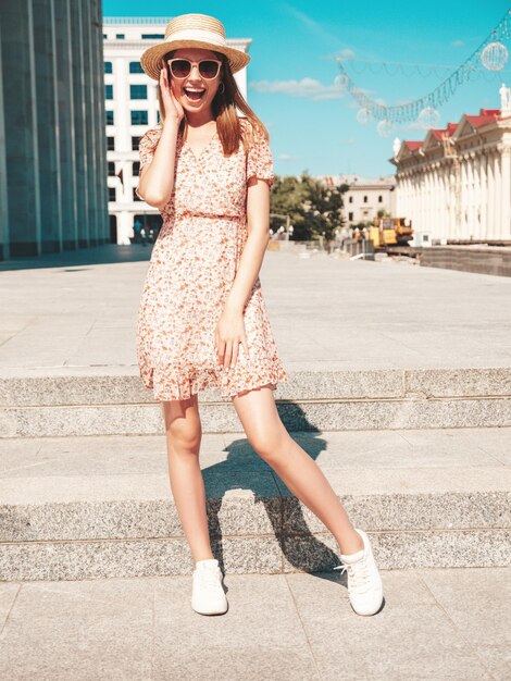 Joven hermosa mujer hipster sonriente en ropa de verano de moda Mujer sexy despreocupada posando en el fondo de la calle al atardecer Modelo positivo al aire libre Alegre y feliz con gafas de sol y sombrero