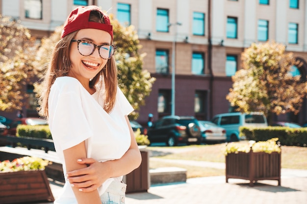 Joven hermosa mujer hipster sonriente en ropa de verano de moda Mujer sexy despreocupada posando en el fondo de la calle con gorra al atardecer Modelo positivo al aire libre Alegre y feliz con gafas de sol