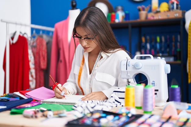 Joven hermosa mujer hispana sastre dibujando en un cuaderno en la fábrica de ropa