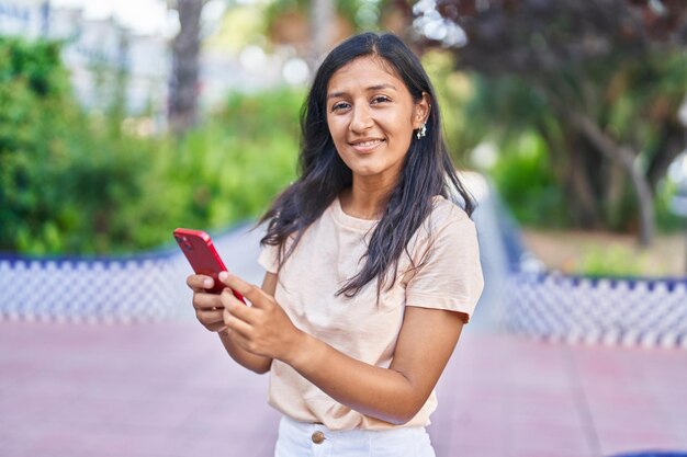 Joven hermosa mujer hispana sonriendo confiada usando un teléfono inteligente en el parque