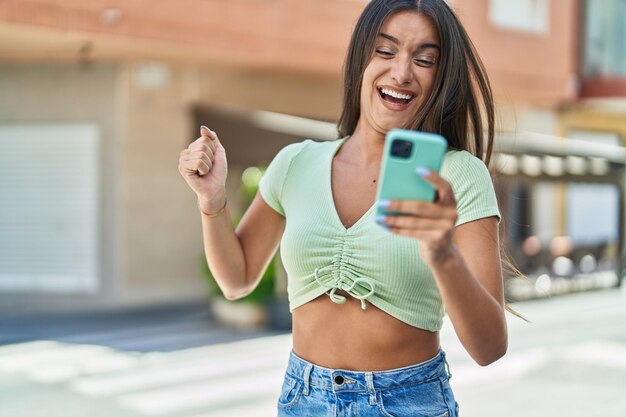 Joven hermosa mujer hispana usando un teléfono inteligente con expresión ganadora en la calle