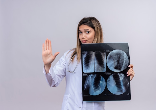 Foto gratuita joven hermosa mujer médico con bata blanca con estetoscopio perforando radiografía de los pulmones haciendo señal de pare con la mano abierta con cara seria con expresión de miedo